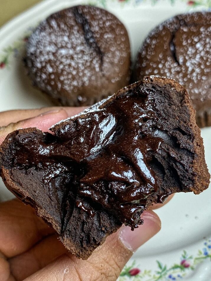 Close-up of a hand holding a decadent chocolate lava cake with a gooey molten center, with more lava cakes in the background.