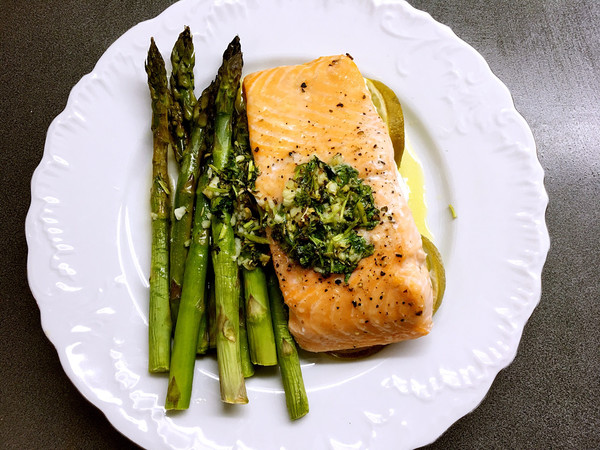 A close-up photo of a flaky, baked salmon fillet drizzled with lemon sauce and topped with fresh asparagus spears on a white plate