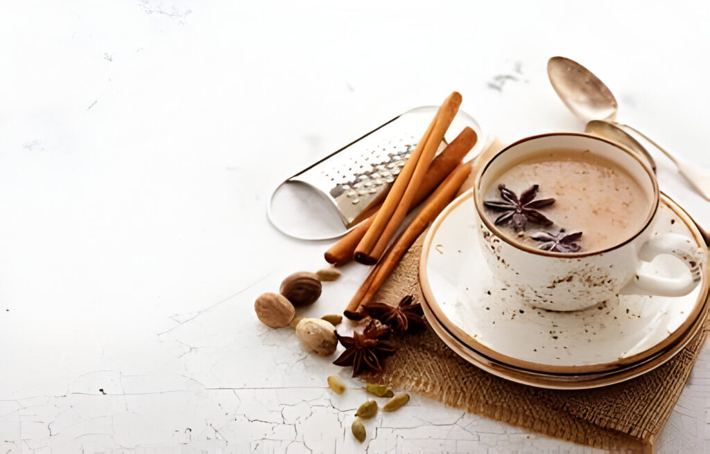 A cup of steaming chai tea on a table with a cinnamon stick and cardamom pods beside it.