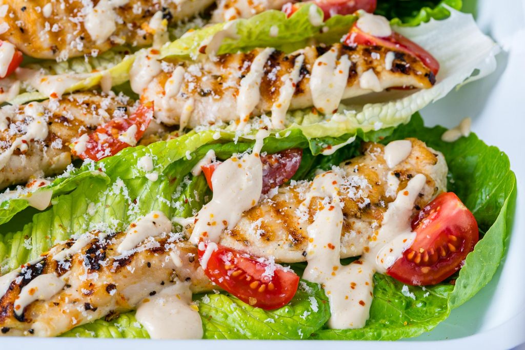 A close-up photo of a halved grilled chicken Caesar wrap on a plate. The wrap is filled with romaine lettuce, grilled chicken, cherry tomatoes, and Caesar salad dressing.