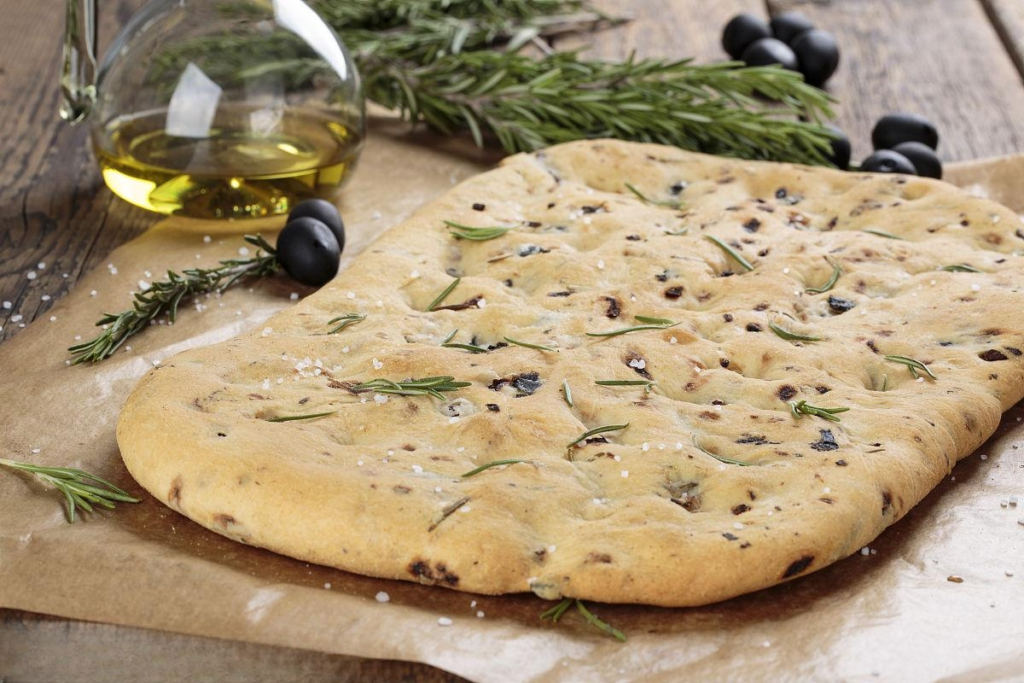 A close-up photo of a round focaccia bread on a table. The bread is golden brown and has dimples filled with olive oil and topped with fresh rosemary sprigs.