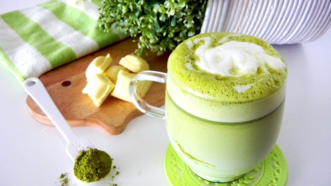 A cup of matcha green tea latte with frothy milk and white chocolate shavings on a table.