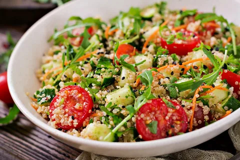 A close-up photo of a colorful quinoa salad on a plate. The salad is made with fluffy quinoa, chopped grilled vegetables (peppers, onions, zucchini), cherry tomatoes, black beans, and fresh cilantro. It is drizzled with a light vinaigrette dressing.