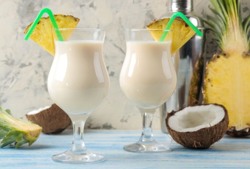 Two glasses of virgin piña colada garnished with pineapple slices, with fresh pineapple and coconut in the background.