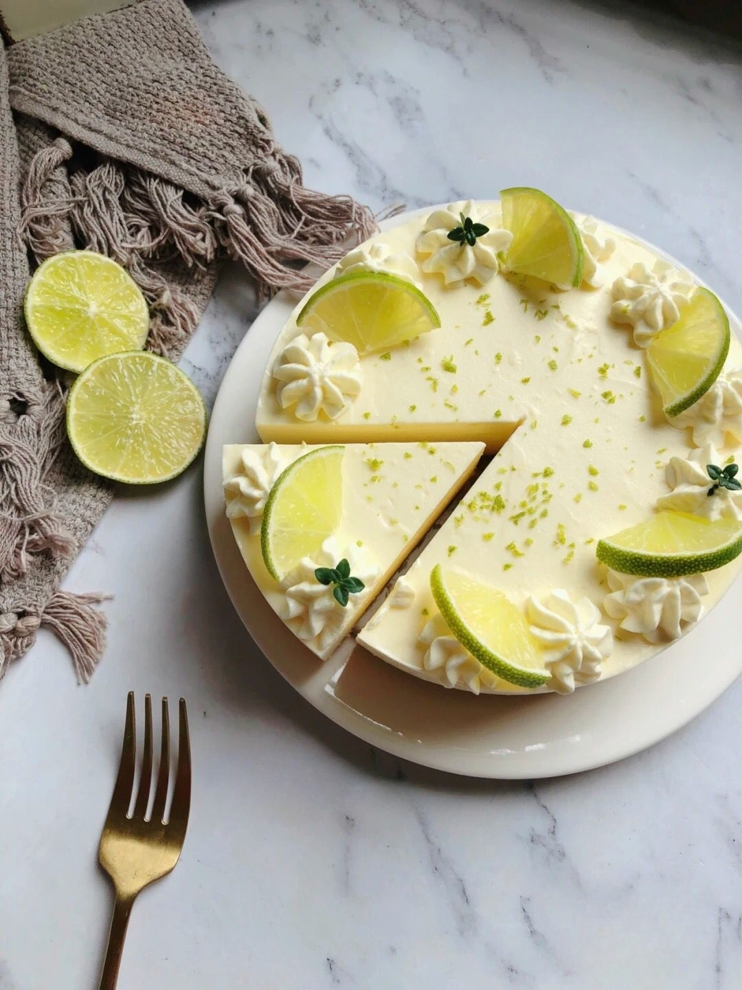 A no-bake lemon cheesecake decorated with lemon slices, whipped cream, and lime zest, placed on a marble surface with a golden fork.