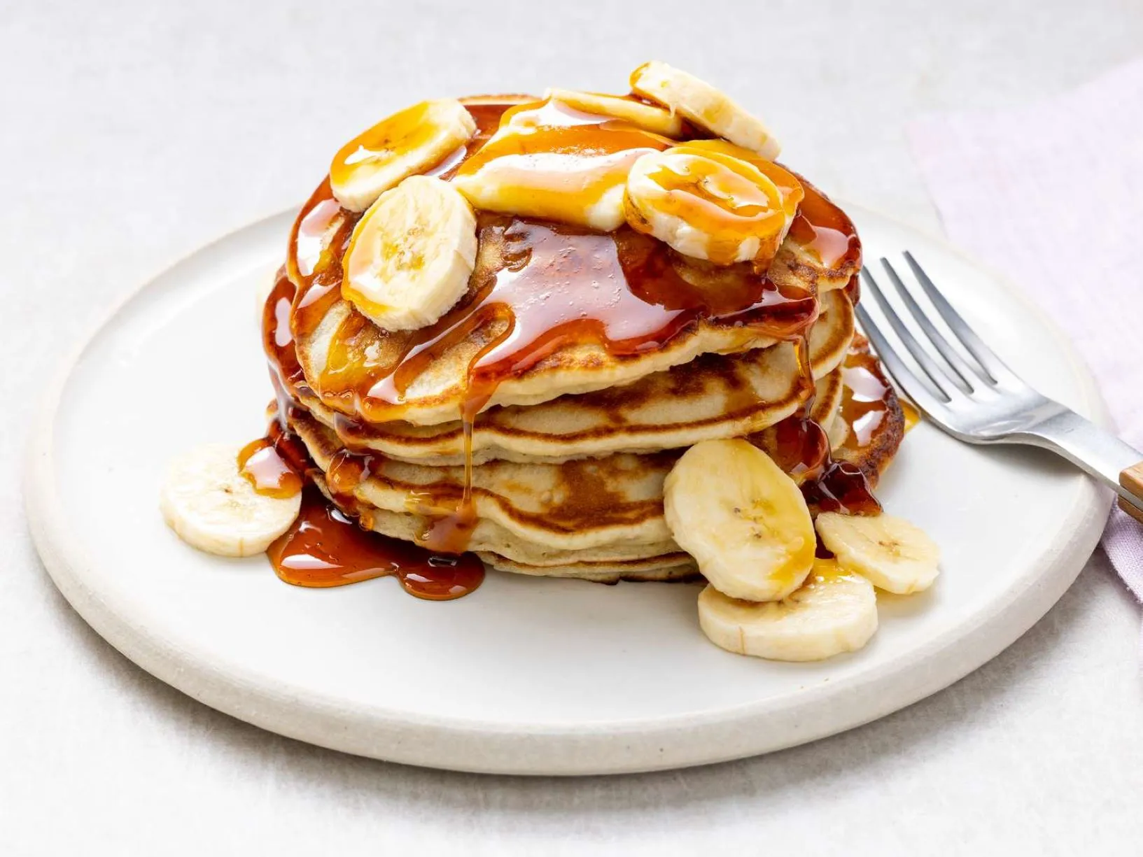 A stack of fluffy pancakes topped with banana slices and walnuts, drizzled with syrup on a white plate.