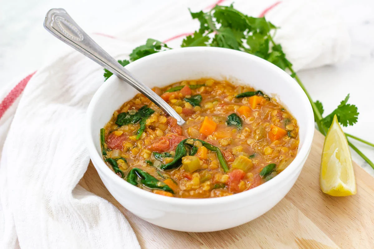 A steaming bowl of lentil soup with carrots, spinach, and a spoon resting on the rim.