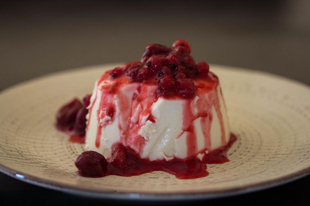 A plate of classic Italian panna cotta topped with fresh raspberries, blueberries, mint leaves, and surrounded by berry sauce.