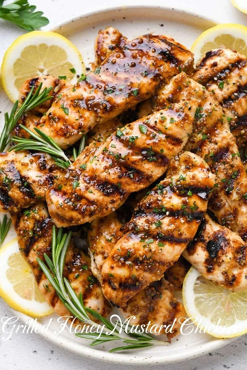 Overhead view of grilled honey mustard chicken breasts on a speckled ceramic plate, garnished with fresh herbs and lemon slices.