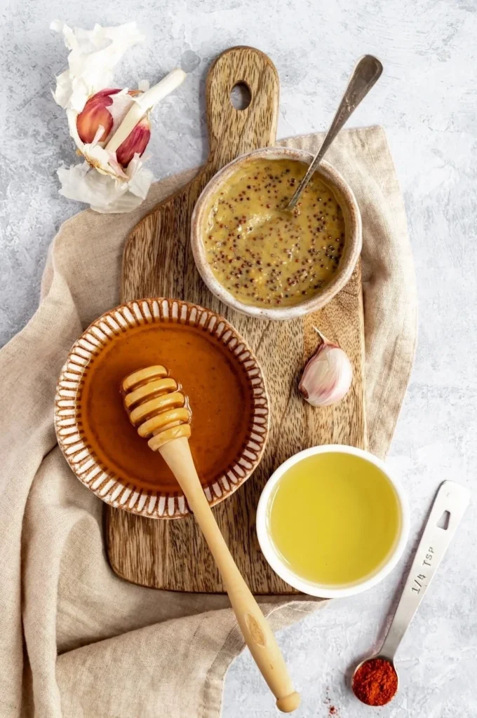  Ingredients for honey mustard grilled chicken including honey, Dijon mustard, garlic, lemon, and spices laid out on a wooden cutting board.
