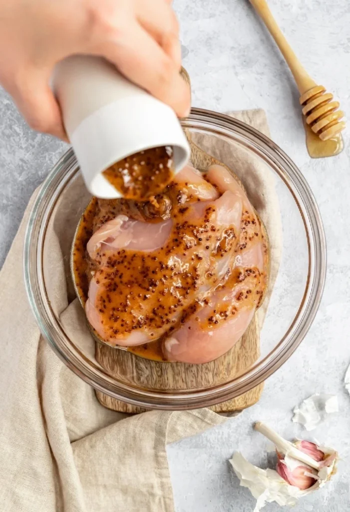 Raw chicken breasts marinated in a glass jar of honey mustard sauce. A hand holds the jar lid while the chicken marinates.
