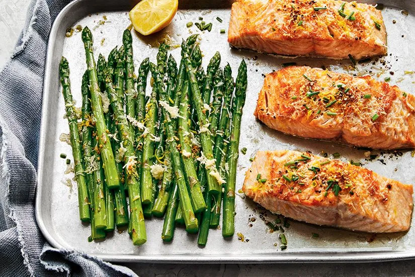 A close-up photo of a sheet pan with roasted asparagus and salmon fillets drizzled with a white sauce.