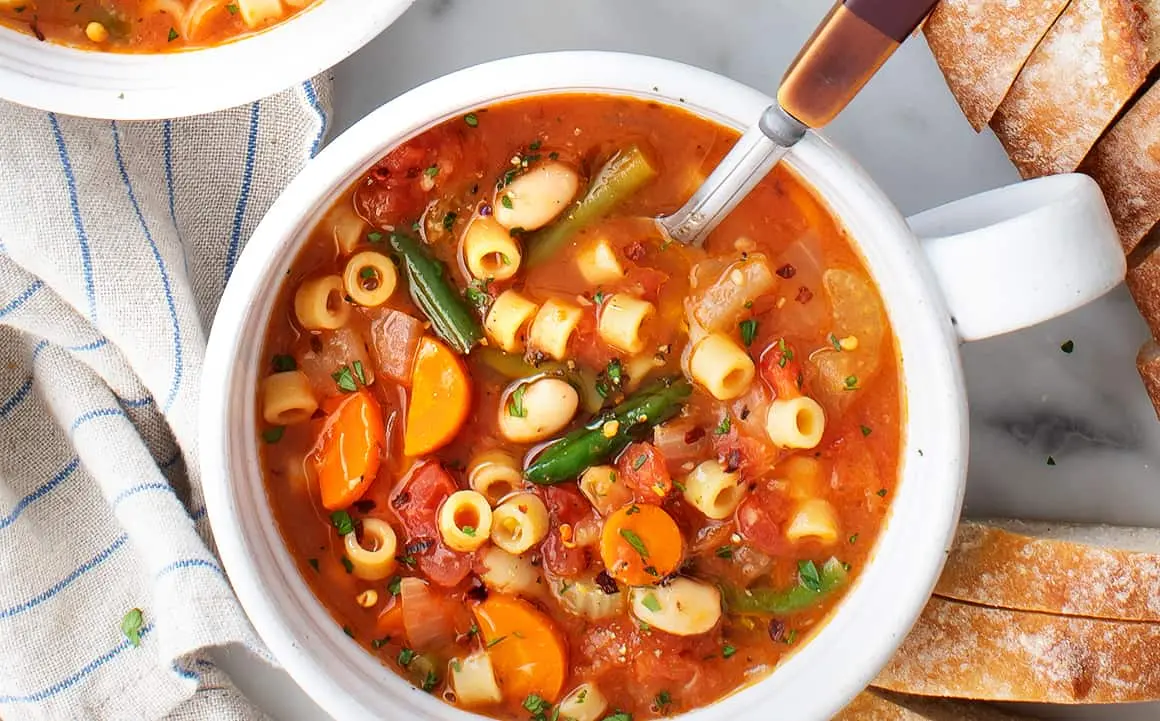 A bowl of hearty minestrone soup filled with fresh vegetables, pasta, and beans, garnished with herbs, and served with slices of crusty bread.