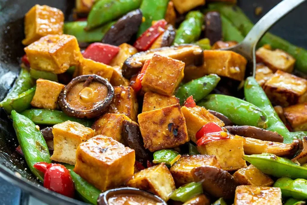 A colorful stir-fry featuring cubes of golden-brown tofu, vibrant green snap peas, red bell peppers, and shiitake mushrooms, all glistening with a savory sauce.