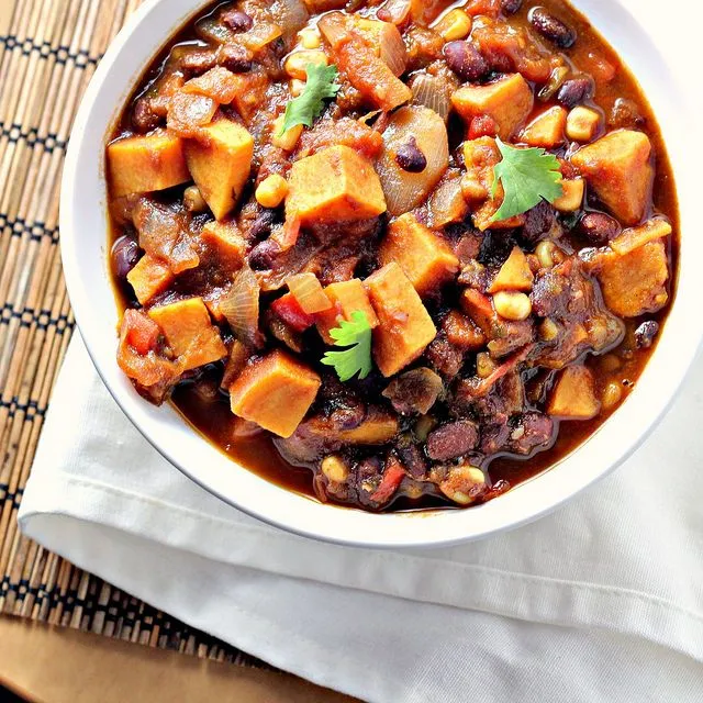 A bowl filled with hearty chili with black beans, sweet potato chunks, corn, and tomatoes.