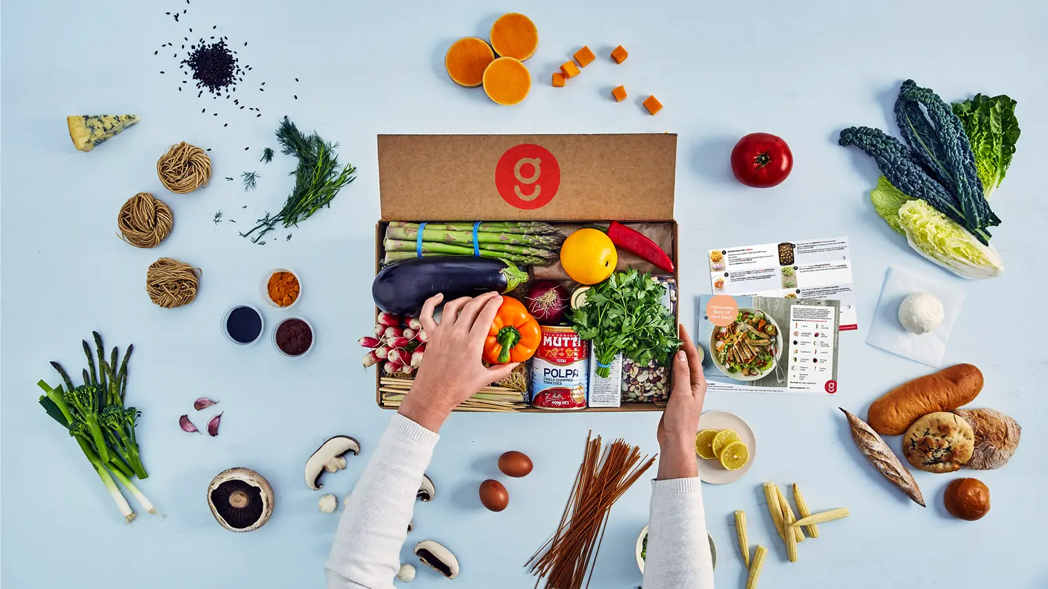 A top-down view of a person organizing a meal kit box with various fresh ingredients and recipe cards on a light blue background.