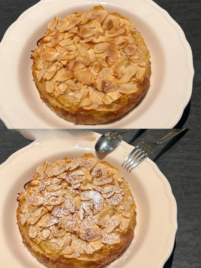 Two golden almond tarts on white plates, one dusted with powdered sugar, served with silverware on a dark surface