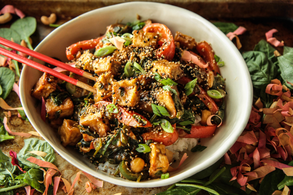 A bowl of Garlic Ginger Tofu Stir-Fry with colorful vegetables, topped with sesame seeds and green onions, served over rice with chopsticks.