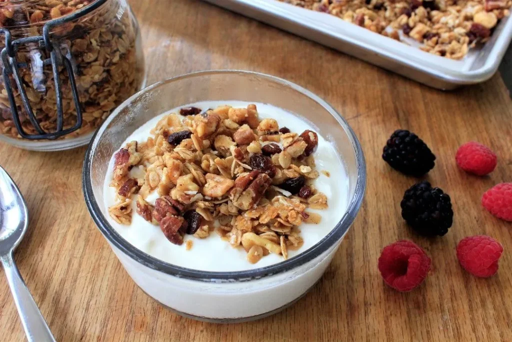 A glass bowl filled with creamy yogurt topped with crunchy homemade granola and surrounded by fresh raspberries and blackberries