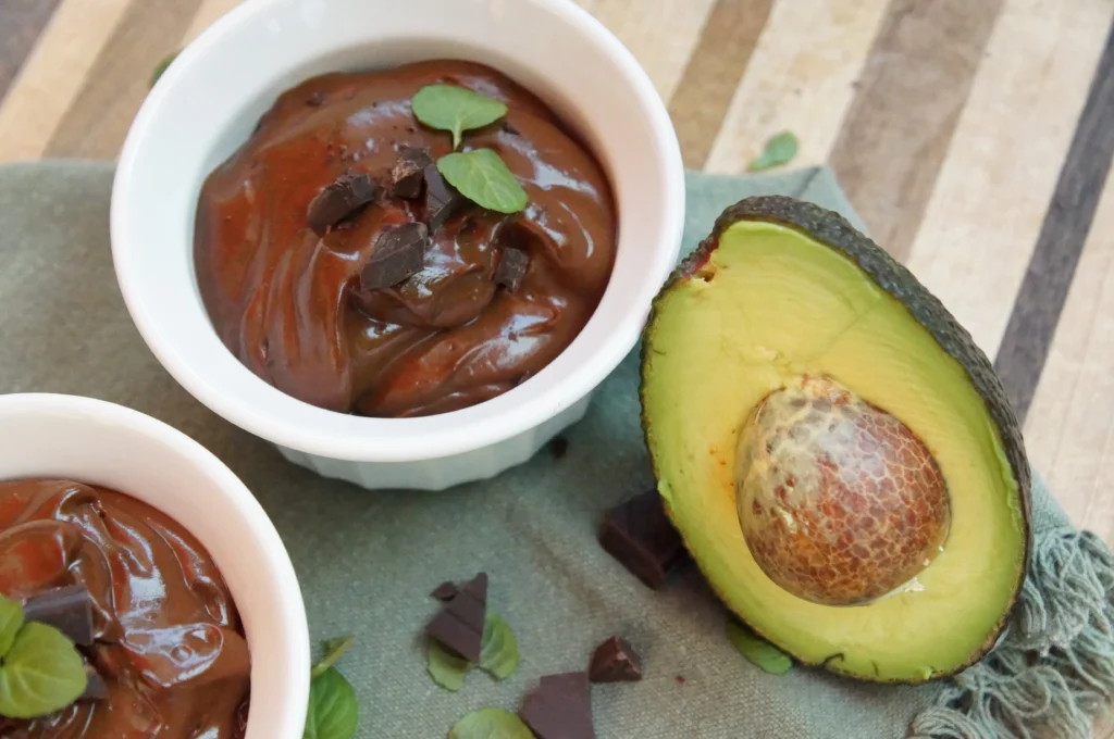 Two bowls of creamy chocolate avocado pudding garnished with chocolate chips and mint leaves, next to a halved ripe avocado