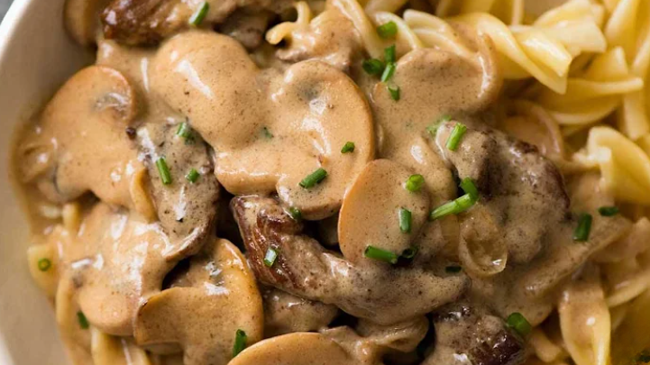 A close-up of a plate of Beef Stroganoff, featuring tender beef slices in a creamy mushroom sauce, garnished with chopped chives and served over egg noodles.