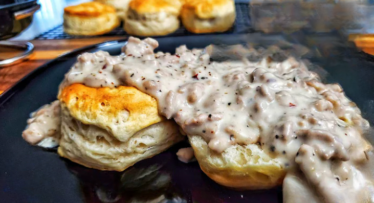 Freshly baked biscuits topped with creamy sausage gravy, served on a black plate with more biscuits in the background.