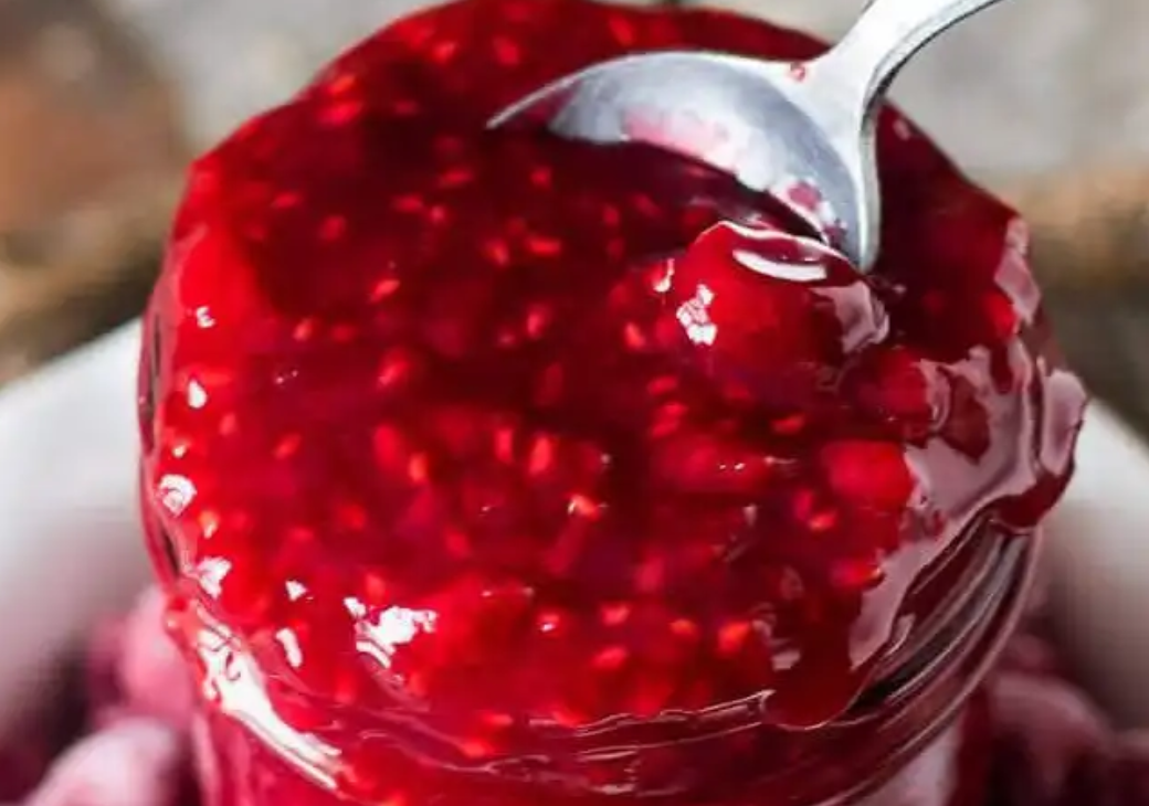 A jar filled with vibrant, homemade raspberry sauce being scooped with a spoon, showcasing its rich, thick texture.