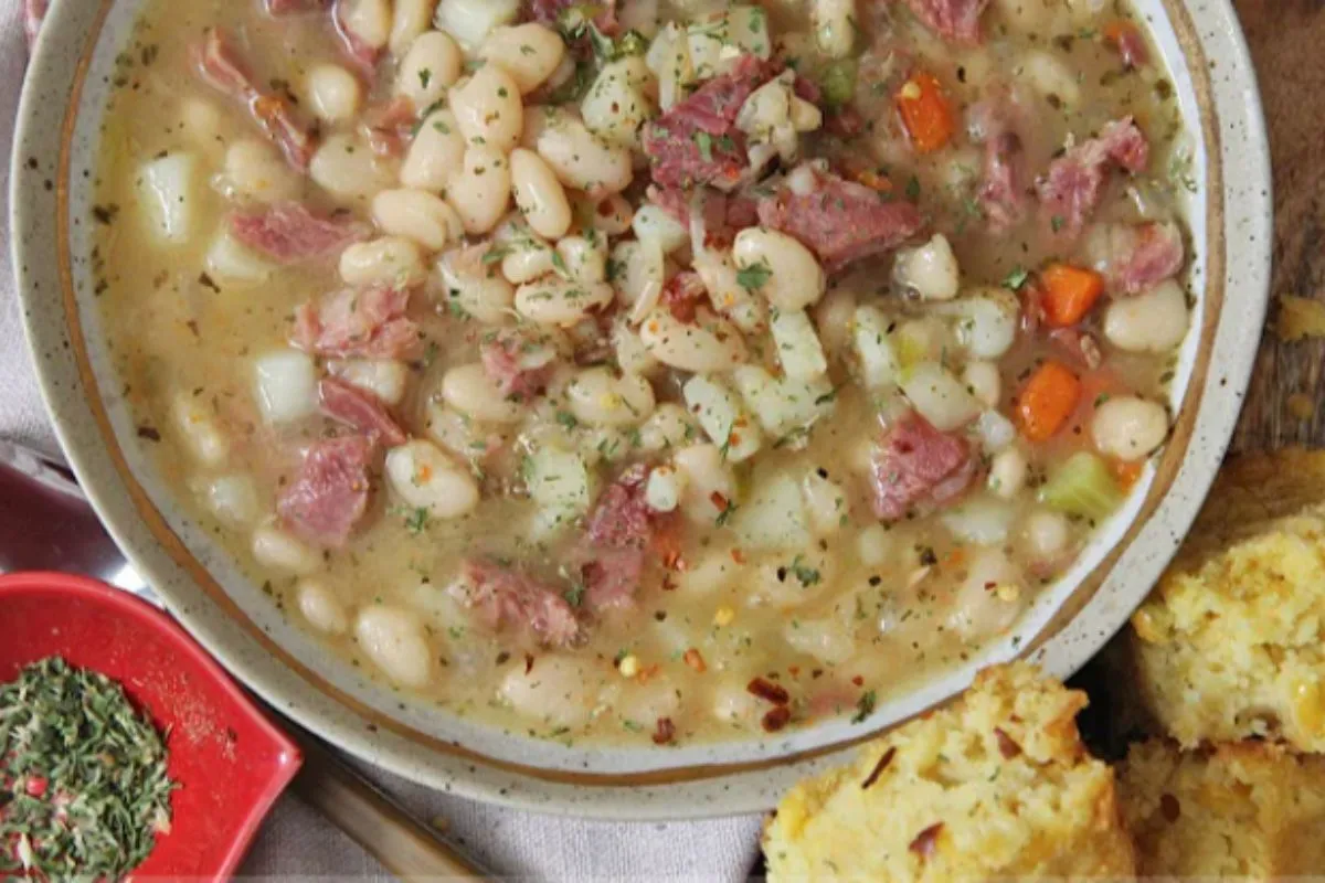 Creamy ham and bean soup in a white bowl, filled with tender white beans, chunks of ham, and colorful vegetables, served alongside golden cornbread muffins