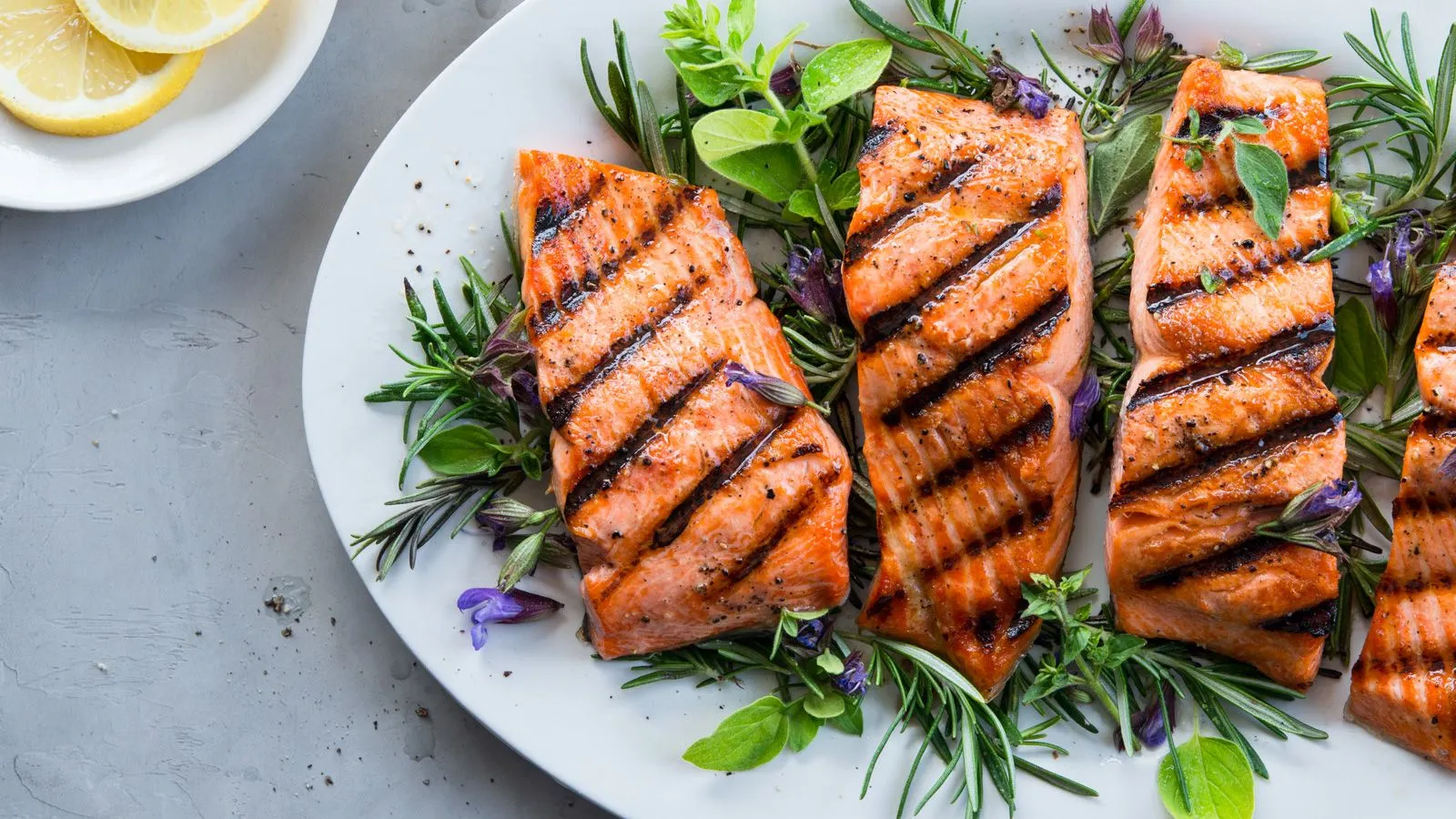 Grilled salmon fillets with herb garnish on a white plate, accompanied by lemon slices.