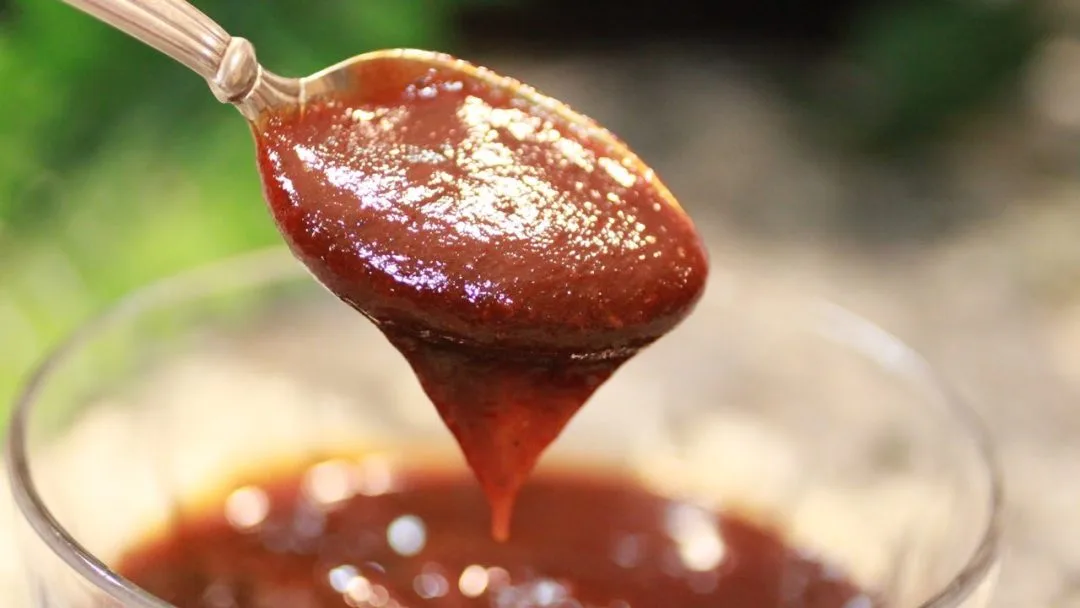 A close-up of a spoonful of rich, homemade BBQ sauce dripping into a bowl.