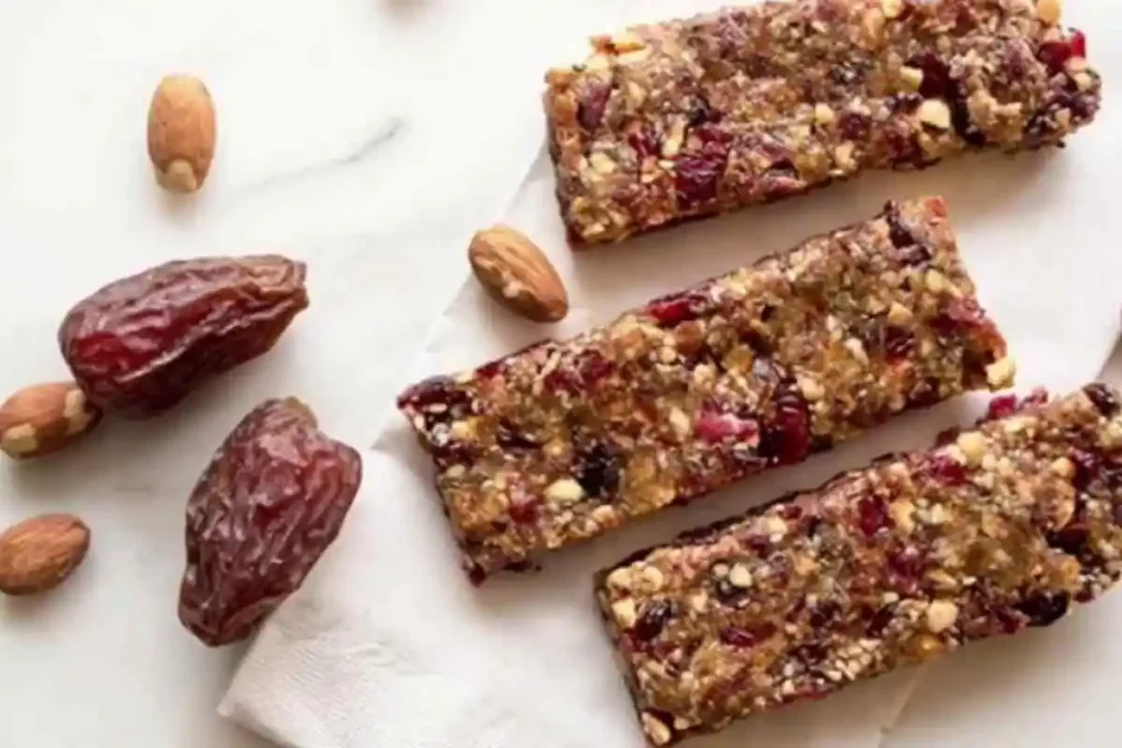 Three homemade date and nut energy bars on a white napkin with whole dates and almonds scattered around.