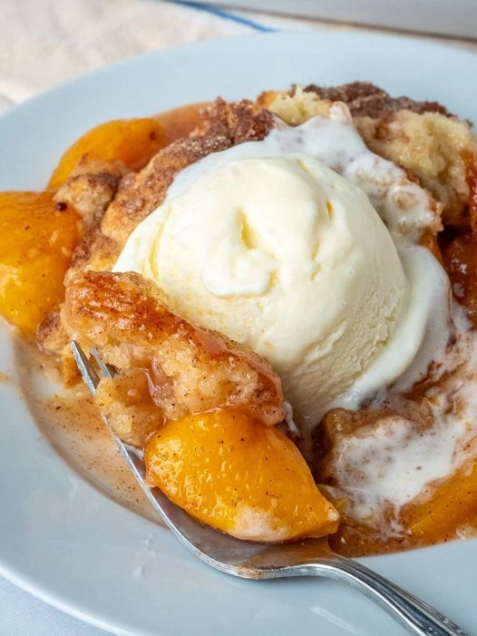 A serving of warm Southern peach cobbler topped with a scoop of vanilla ice cream on a white plate