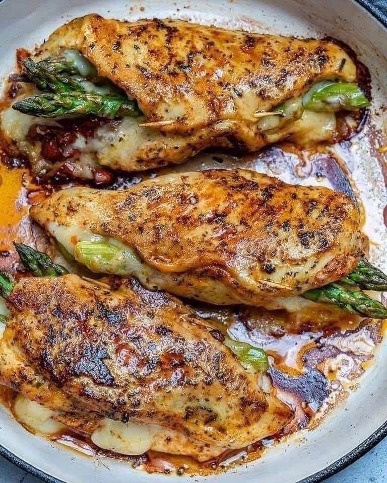 Three golden-brown asparagus stuffed chicken breasts in a skillet, showing melted cheese and green asparagus tips