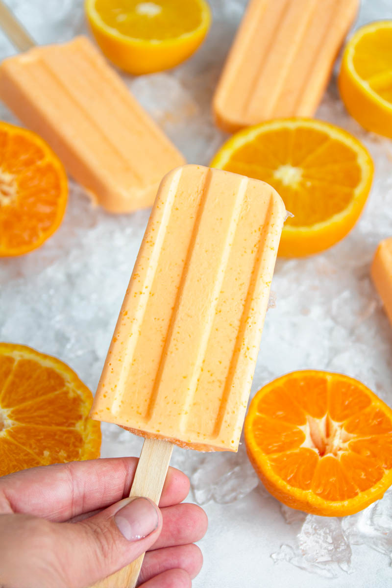 Hand holding a creamy orange popsicle surrounded by fresh orange slices and other popsicles