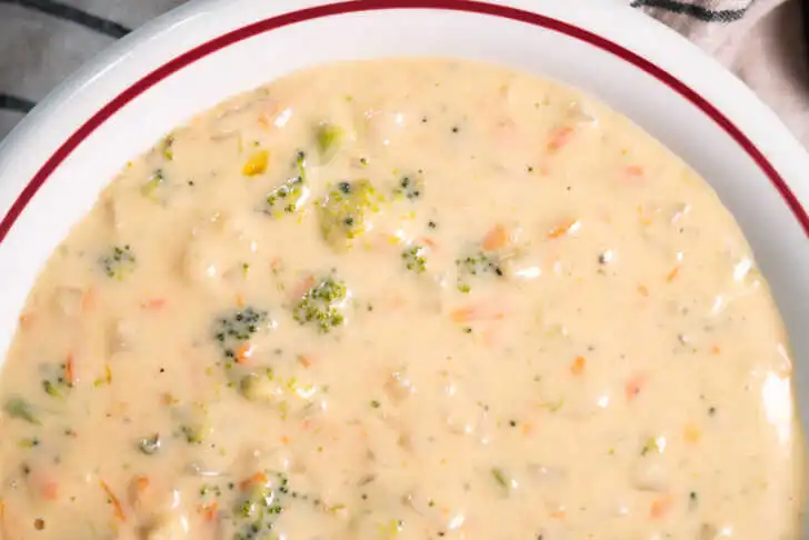 Close-up of thick, creamy broccoli cauliflower cheese soup with visible vegetable pieces in a white bowl with red rim