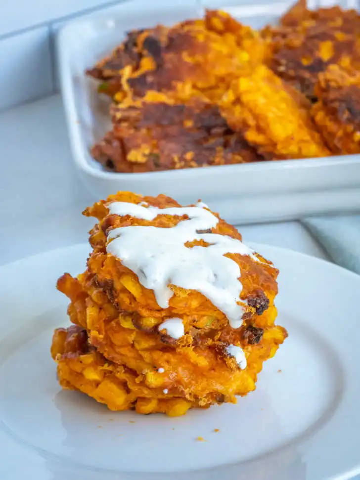 Crispy golden corn fritters stacked on a white plate, topped with creamy sauce, with more fritters in a baking dish in the background