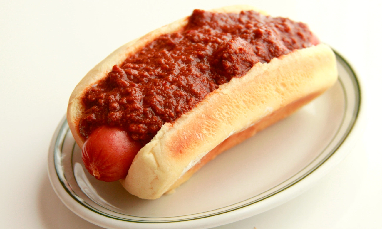 A chili dog on a white plate, featuring a grilled hot dog in a soft bun topped with generous amounts of meaty red chili sauce