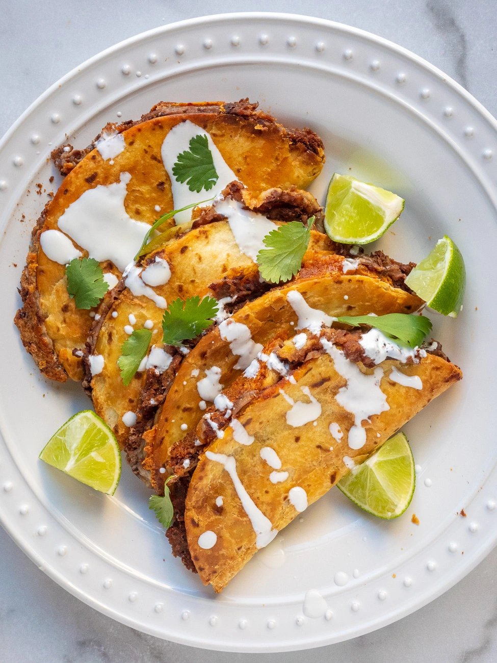 Crispy black bean tacos on a white plate, topped with sour cream, cilantro, and served with lime wedges