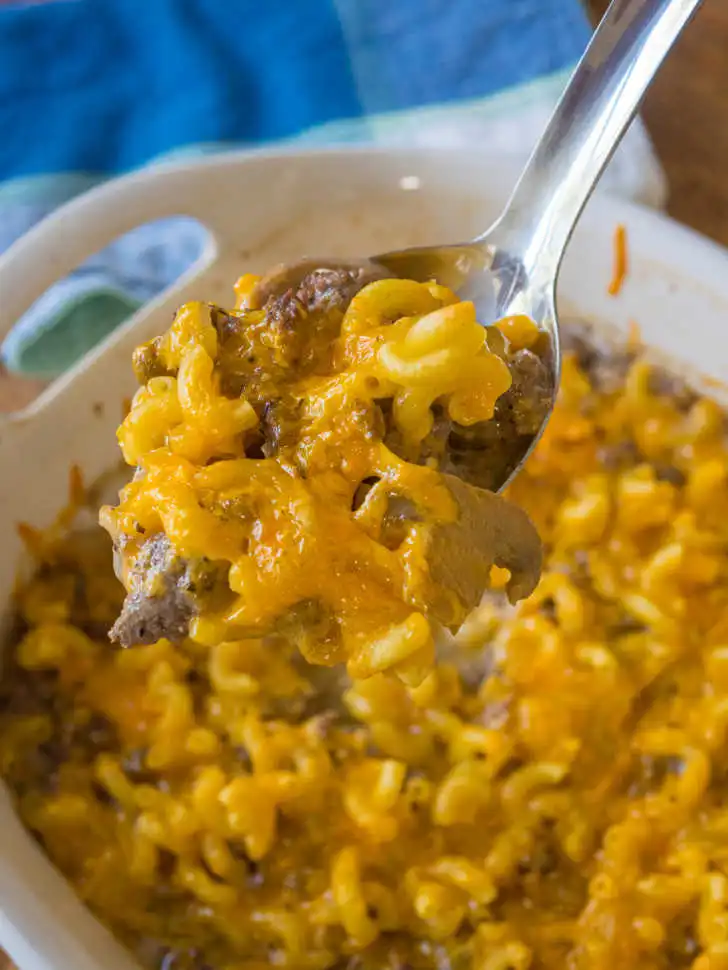 Close-up of a spoon lifting a serving of Hamburger Supreme Casserole, showing gooey melted cheddar cheese stretching over tender macaroni and savory ground beef.