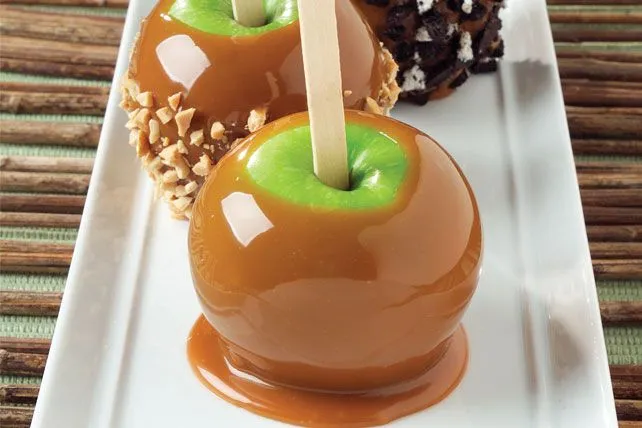 Two glossy caramel-coated apples with chopped nuts on a white plate, next to chocolate-covered treats